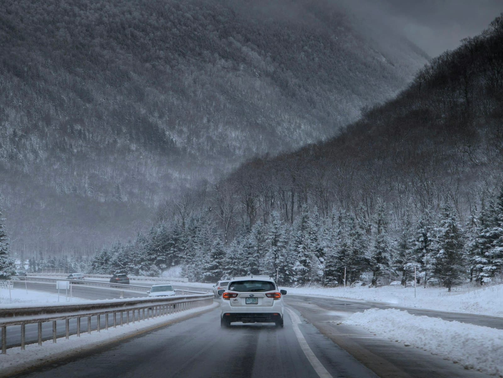 Car on a snow-covered mountain highway during active snowfall, guardrails lining the road